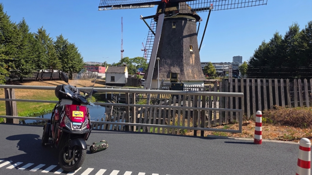 De rode Tukkie scooter geparkeerd op een bruggetje met een traditionele rietgedekte molen op de achtergrond in de regio Den Haag.