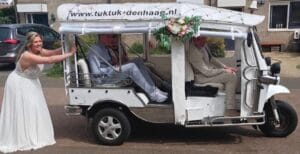 Laughing bride pushing the white wedding tuk tuk with flowers, guests watching, in The Hague